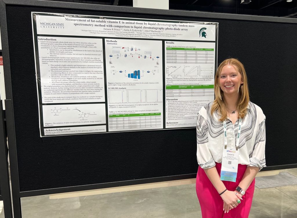 Woman in white and black blouse and pink pants stands in front of her research poster.